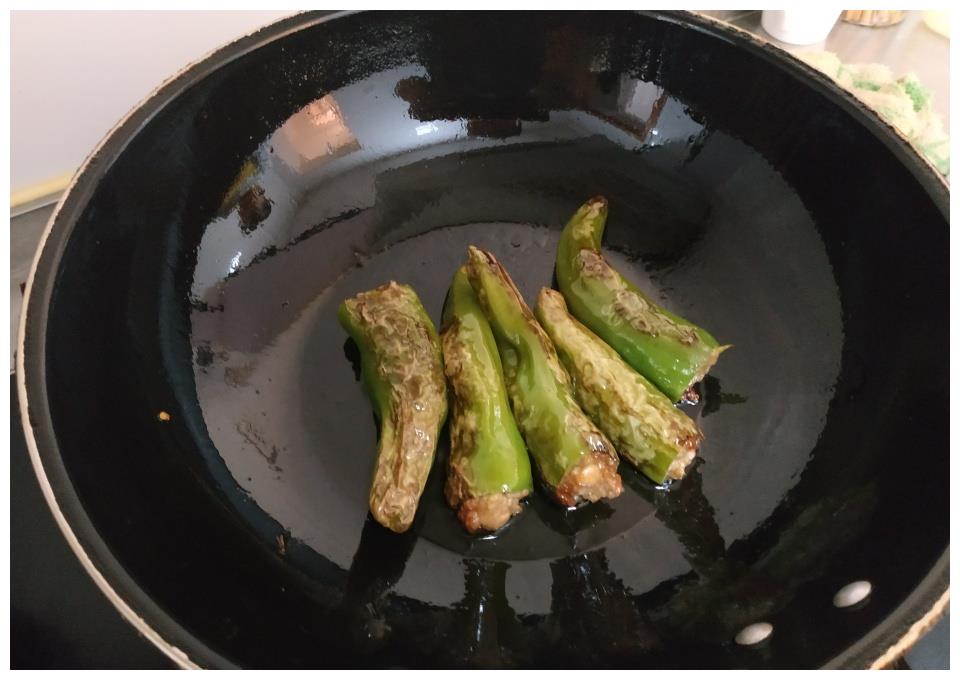 一根根青绿色尖椒在黑色不粘锅中被压扁,表面已呈现出明显的深褐色焦斑,形成类似老虎皮毛的纹理,锅边配有木铲和调料碗,展现传统中式烹饪技法 虎皮尖椒煎制过程