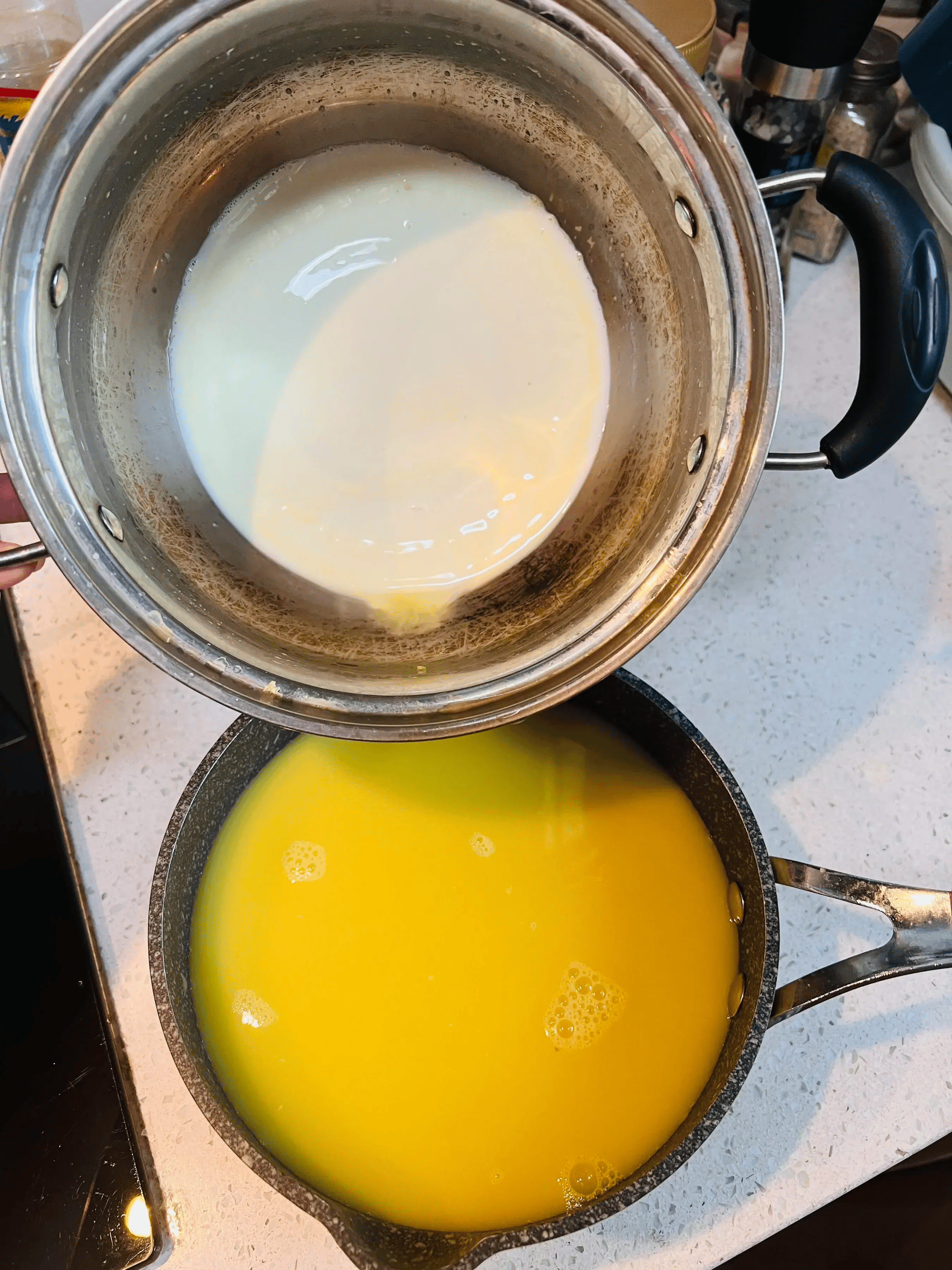 一个不锈钢奶锅置于炉灶上,锅内为微微冒泡的乳白色液体,一只手正拿着木勺顺时针搅拌;旁边台面上摆放着量杯、橙汁、淡奶油瓶和糖罐,整体呈现专业厨房操作场景 奶基混合与加热过程