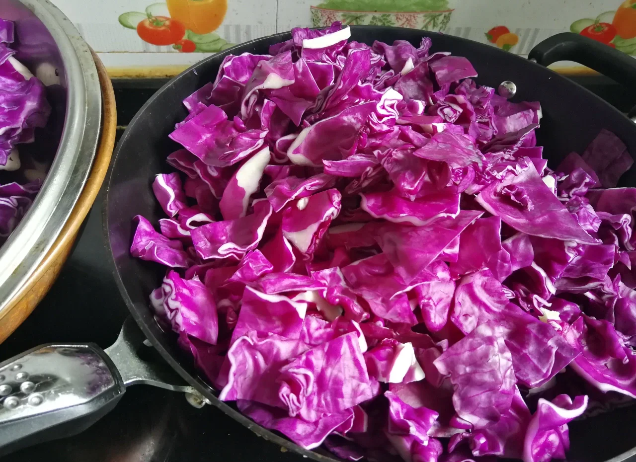 左侧为切丝后的紫甘蓝用于制作凉拌菜,颜色鲜艳;右侧为锅中翻炒的紫甘蓝,边缘微焦,配有蒜末和橄榄油,旁边放置木铲和调料瓶,展示不同烹饪方式 紫甘蓝凉拌与炒菜对比图