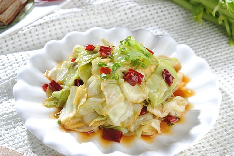 厨房场景：不锈钢灶台上正在翻炒的新鲜包菜丝，热油轻溅，包菜呈半透明翠绿色，边缘微卷，配蒜末与红椒丝点缀，锅气升腾可见细微水汽