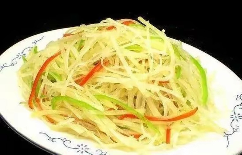 一盘装在白色瓷盘中的清炒土豆丝，金黄色的细丝整齐排列，点缀少许红辣椒丝和葱段，背景为木质餐桌，光线柔和自然