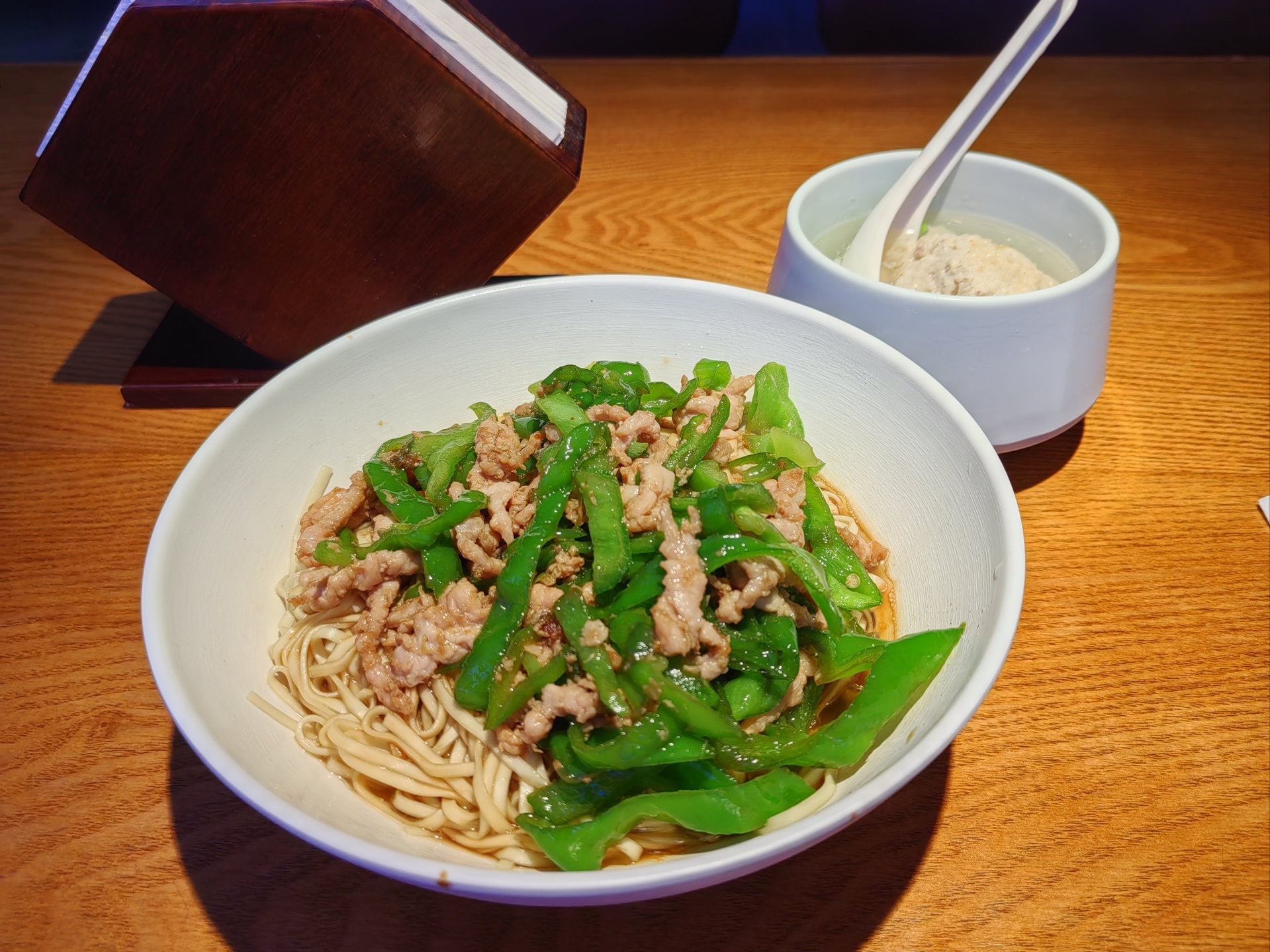 一碗热气腾腾的椒江姜汤面摆放在木质餐桌上,汤色微黄透亮,上面覆盖着金黄的荷包蛋、翠绿青菜、粉色瘦肉片和虾干,面条整齐卷曲,周围点缀少许葱花,整体呈现温暖诱人的视觉效果 椒江姜汤面成品全景图
