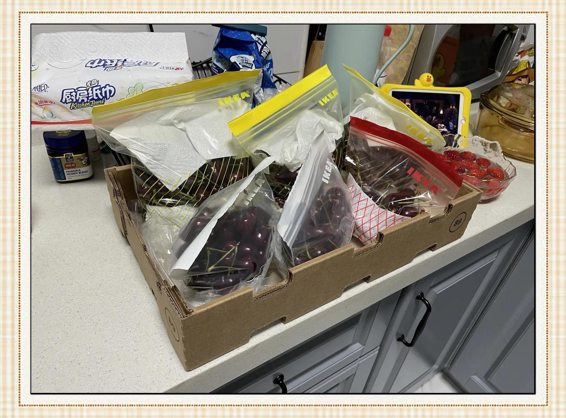 透明塑料盒内整齐摆放着带绿蒂的新鲜红草莓，盒底垫有白色纸巾，整体置于浅灰色台面上