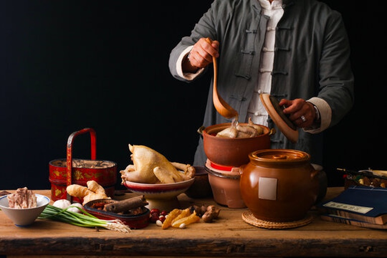 一个中式厨房环境中,一位女性正在用白色陶瓷砂锅炖煮鹿鞭鸡汤,锅中漂浮着鹿鞭片、鸡肉块、枸杞和姜片,热气腾腾,旁边摆放着装有干鹿鞭片的玻璃罐和中药材袋,整体氛围温馨养生 家庭炖煮鹿鞭汤场景