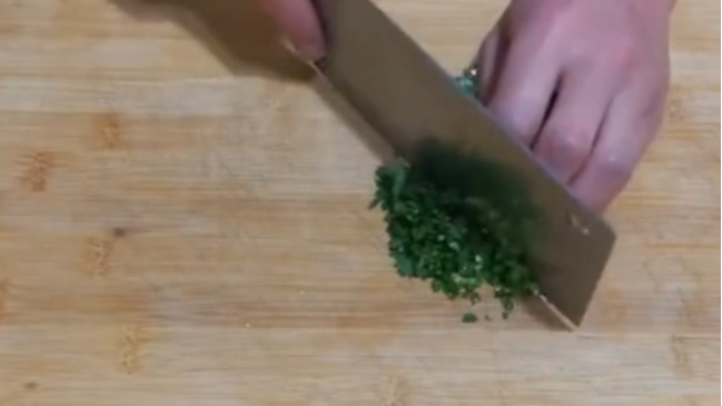一位厨师正在木质砧板上用不锈钢刀具切碎新鲜香菜,刀下是已切好的绿色碎叶,旁边放有陶瓷碗和橄榄油瓶,展现健康料理过程 厨房中切碎香菜的场景