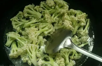 图片说明:一位家庭主妇在明亮厨房中正在将焯过水的西兰花和切丝的紫甘蓝放入炒锅中翻炒,灶台上有酱油瓶、蒜瓣和橄榄油,展现健康家常菜制作过程,突出食材的新鲜与安全性 家庭烹饪场景