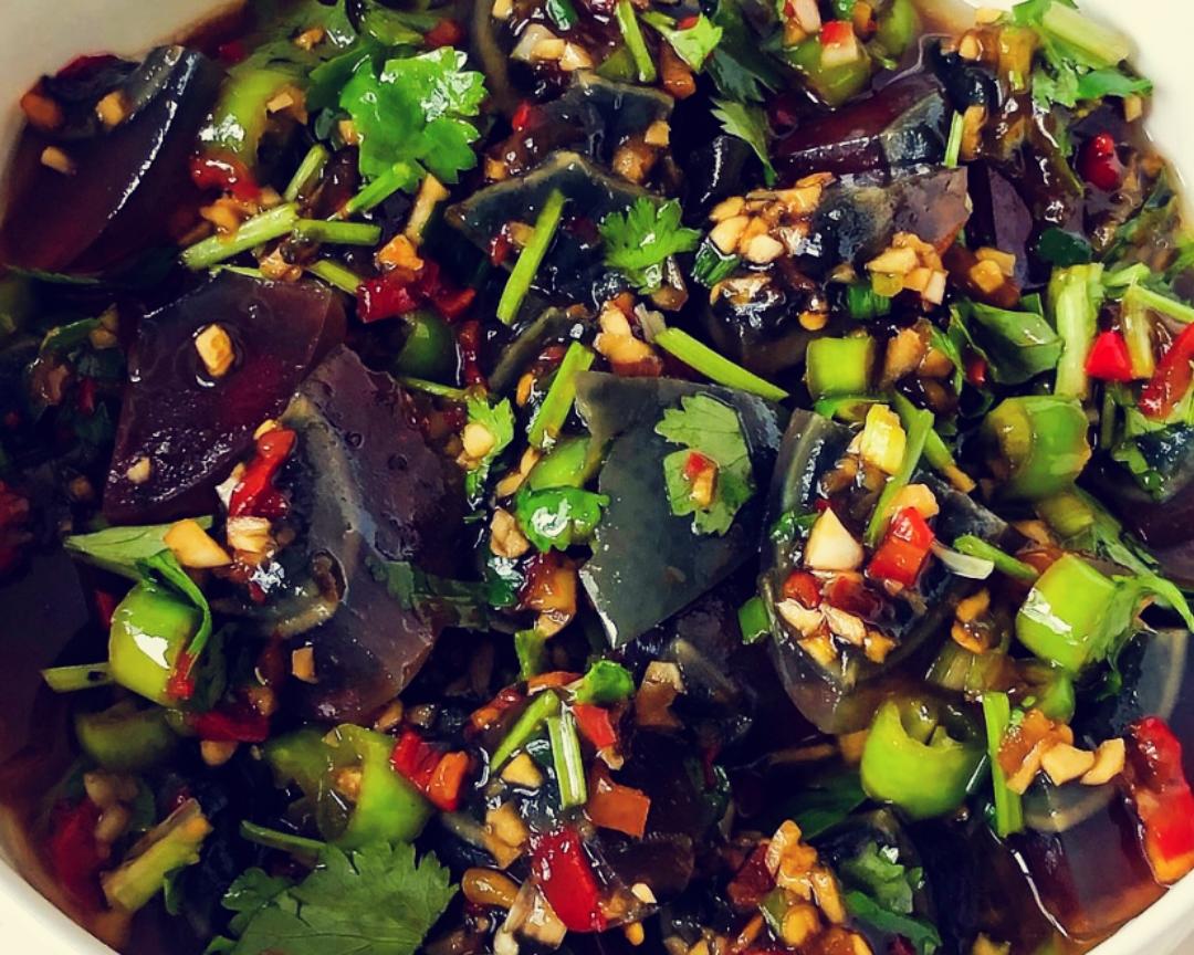 高清俯拍:左侧为3根鲜红牛角椒平铺于竹砧板上,表皮光亮无瑕;右侧为2颗剥壳皮蛋浸在透明玻璃碗的浅层淡盐水中,水面浮着两片姜片,背景虚化突出食材新鲜度 红椒与皮蛋预处理特写
