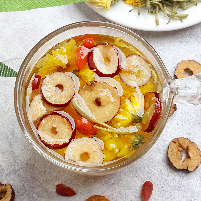 一个透明玻璃杯中盛着金黄色的茶汤,一朵完全盛开的金丝皇菊居中漂浮,周围点缀几颗红色枸杞,杯旁放有一小碟枸杞和一朵干菊,背景为原木茶桌与绿植 金丝皇菊与枸杞同泡的玻璃杯