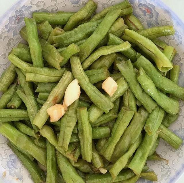 无筋豆快炒出锅特写：青翠豆段整齐铺于白瓷盘中，表面油润光亮，散落金黄蒜末与微红椒段，背景虚化显示燃气灶蓝色火苗正旺