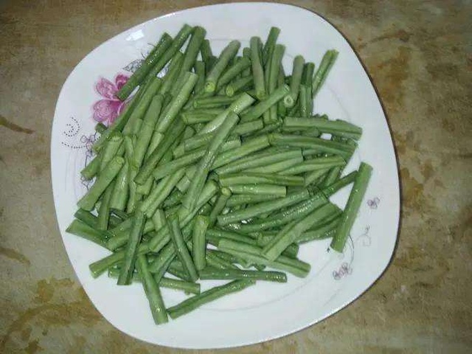 一位女性在明亮厨房中操作,身穿围裙,手持刀具正在处理新鲜绿色豆角,动作清晰展示切除顶端和底端的过程,豆角整齐排列在白色瓷盘中,背景可见灶台和锅具 家庭厨房场景,一位主妇正在砧板上切除豆角两头并撕去侧边筋络,旁边放着一盘处理好的豆角段