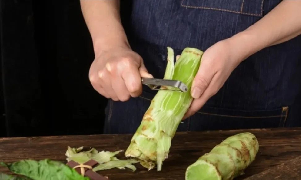 案板上摆放着已去皮的莴苣段,一把锋利的厨师刀正在将其纵向切开,旁边有一把小勺正从莴苣内部刮除白色髓芯,另一侧是切好的均匀莴苣丝堆叠在白色瓷碗中 莴苣切丝与去芯过程