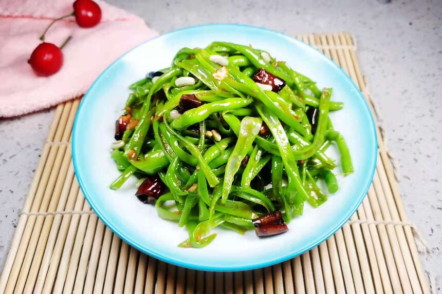 四季豆健康搭配场景：干煸四季豆盛于浅口陶盘，左侧配一小碗紫薯糙米饭，右侧放三颗烤香的金黄松子仁，背景虚化处可见新鲜番茄与西兰花