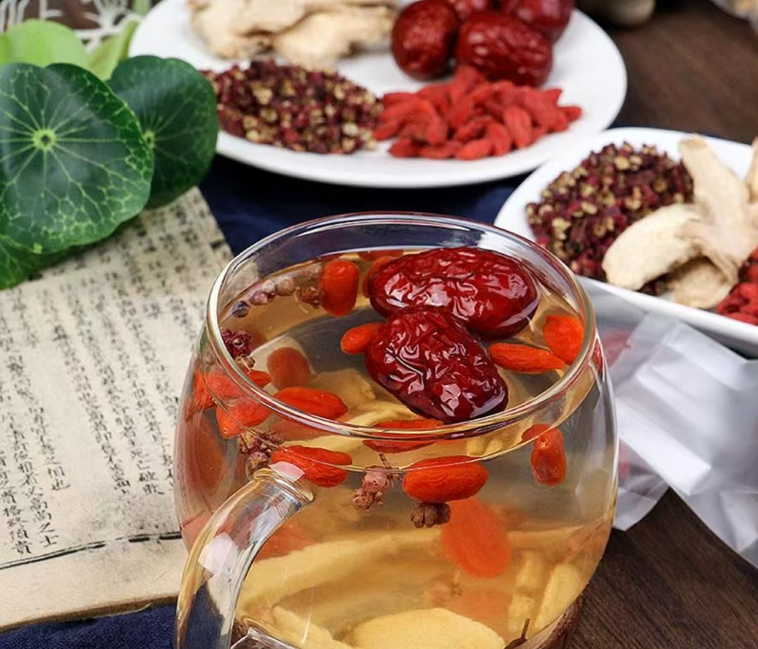 透明玻璃茶杯中热水冲泡的红枣桂圆枸杞茶,可见红枣切片、完整桂圆仁和悬浮的枸杞颗粒,水面微泛金红色光泽,旁边放置陶瓷茶匙和未使用的干料样品,体现家庭自制场景 玻璃杯中冲泡的红枣桂圆枸杞茶