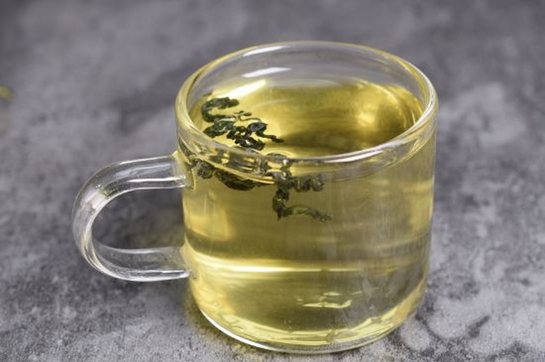 一位女性手持玻璃茶壶正在冲泡丹桂叶茶,透明茶汤呈现淡黄色,叶片在水中缓缓舒展,旁边放置干燥的丹桂叶样品和检测报告复印件,桌面为木质纹理,环境整洁明亮 丹桂叶茶冲泡过程展示