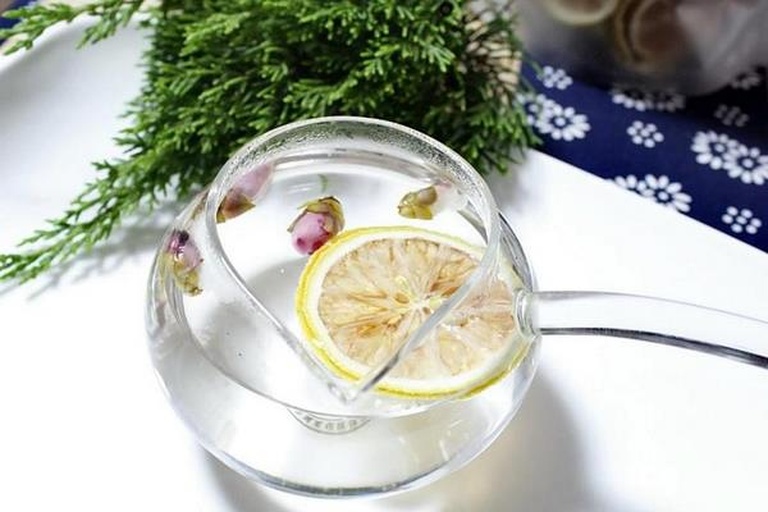 一位女性的手正端着一杯透明玻璃杯装的金桔柠檬茶,杯中可见清晰的金桔片和柠檬片漂浮在淡黄色液体中,杯壁凝结水珠,背景为明亮的窗台,阳光洒入,呈现出清新自然的生活场景 手握玻璃杯中的金桔柠檬茶