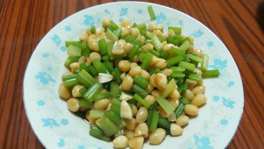 一位厨师在家庭厨房灶台前使用铁锅翻炒蚕豆与芹菜混合菜肴,锅中食材呈翠绿色,热气升腾,旁边配有蒜瓣、盐罐等调料,体现日常健康烹饪情境 家庭厨房中正在翻炒蚕豆与芹菜的场景