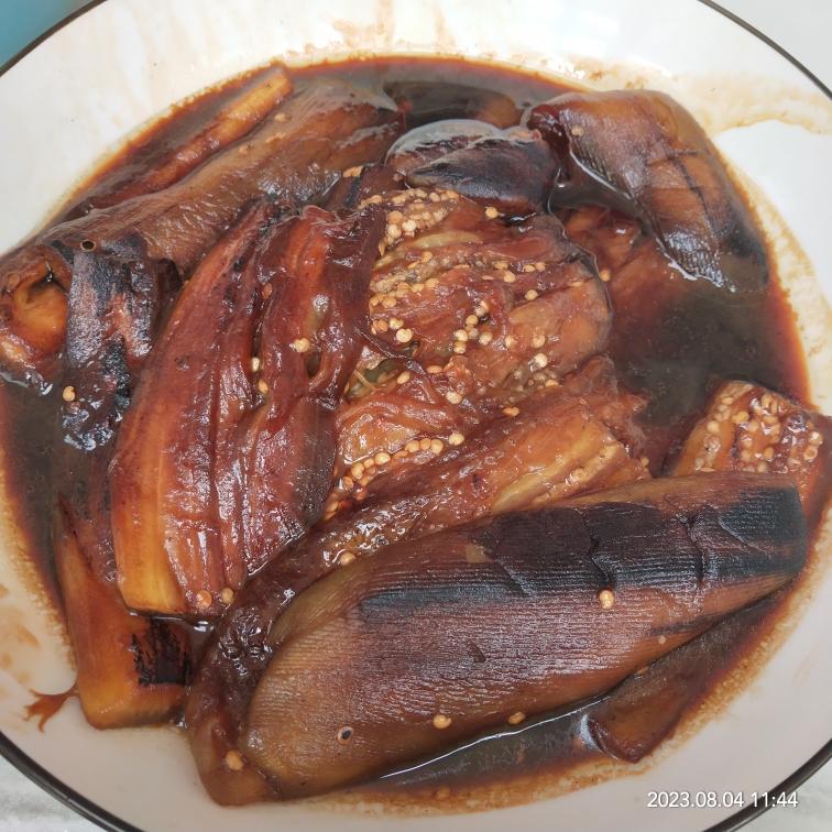 一盘刚出锅的不过油红烧茄子盛放在白色瓷盘中,茄条整齐排列,表面裹着浓稠红亮的酱汁,点缀着绿色葱花和红色椒片,背景虚化的餐桌上配有米饭和筷子,展现家常用餐氛围 不过油红烧茄子成品特写