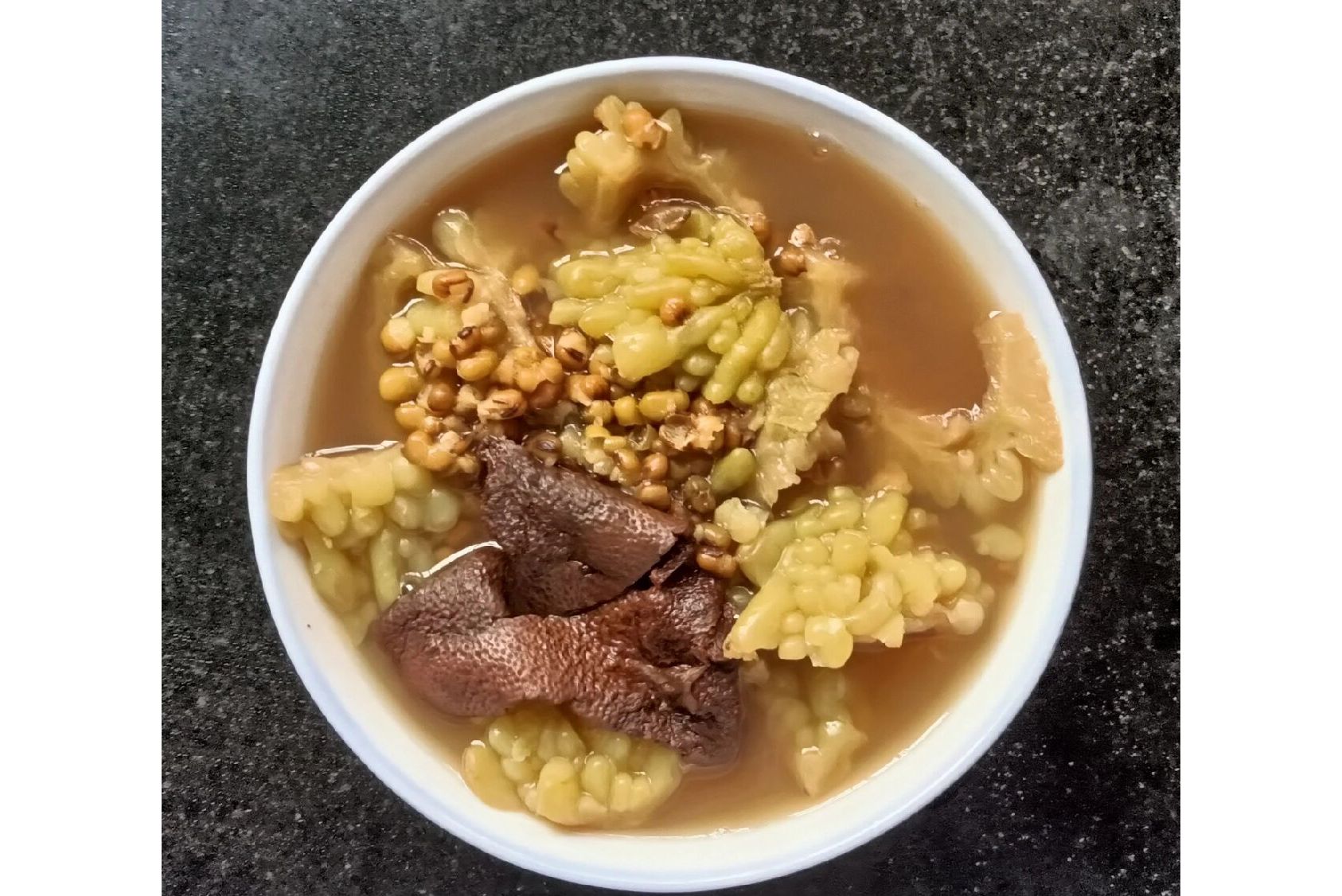 苦瓜绿豆汤成品图：青碧色汤液盛于白瓷碗中，表面浮有细小绿豆皮碎屑，汤色清澈透亮，碗沿搁置一片新鲜苦瓜薄片作点缀，背景为木质餐桌与阳光照射的窗棂