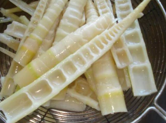 一口不锈钢锅中沸水翻滚,几段去壳切好的乳白色竹笋正在焯水,旁边放着砧板、刀具和剩余的带壳竹笋,展现家庭处理竹笋的标准流程 家庭厨房中正在锅中焯水处理新鲜竹笋的过程