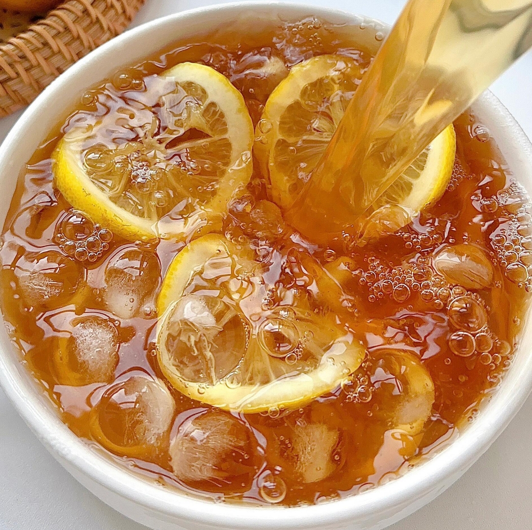 高清特写:成品柠檬冰红茶,汤色清澈透亮呈琥珀金色,可见悬浮的嫩绿柠檬皮丝,杯壁覆满冷凝水珠,背景有新鲜青柠檬横切面展示饱满果肉与油亮表皮 一杯透亮金黄色的柠檬冰红茶特写,杯中悬浮着几缕翠绿柠檬皮丝,杯壁凝结细密水珠,背景为木质料理台与新鲜青柠檬切片
