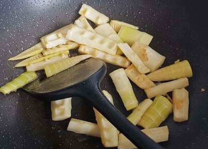 一位家庭主妇在厨房灶台前翻炒锅中的黑竹笋,锅中食材色泽油亮,配有青红椒丝和腊肉片,旁边放置着切好的笋段和调料碗,环境整洁温馨 家庭厨房中正在炒制黑竹笋的场景