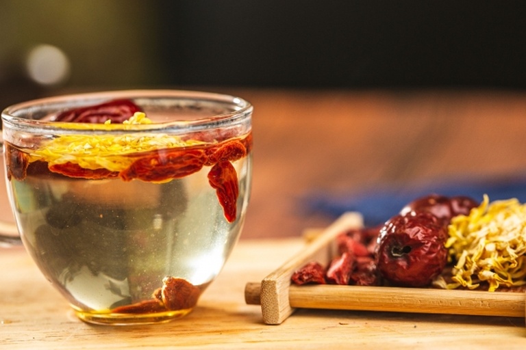 白色瓷杯中盛放一杯刚冲泡好的野菊花陈皮茶,茶汤透亮呈浅琥珀色,杯边放置一小段卷曲的深棕色陈皮和几朵干制野菊花,整体呈现自然健康的饮品氛围 野菊花陈皮茶饮成品展示