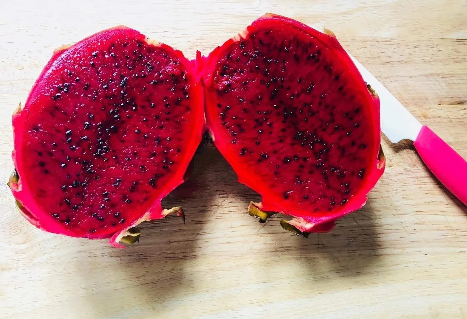 一个成熟的红心火龙果被横向切开,露出鲜艳的紫红色果肉,黑色小籽均匀分布,果汁饱满,果皮呈亮粉色带绿色鳞片,背景为木质砧板,周围散落几片果肉和一片绿叶,光线柔和,突出果实的新鲜与视觉冲击力 红心火龙果切开特写