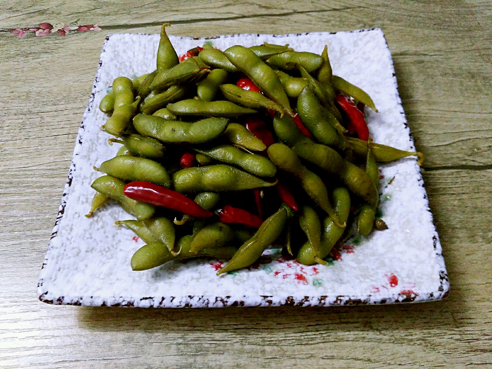 一盘盛放在白色瓷碗中的凉拌毛豆节,颜色翠绿诱人,表面淋有油亮酱汁,点缀蒜末和红辣椒圈,背景为竹制餐桌垫 凉拌毛豆成品图