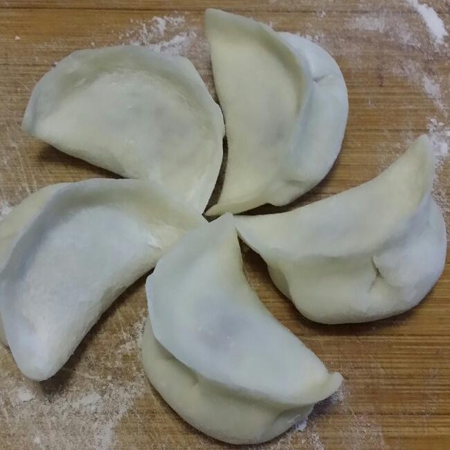 俯拍视角:木质案板上铺薄粉,左手托面剂旋转,右手持擀面杖由中心向边缘推擀,面皮呈均匀圆形,边缘透光可见薄度,旁置已包好的3枚香芹水饺(褶皱清晰、肚圆饱满) 手工擀制香芹水饺面皮过程