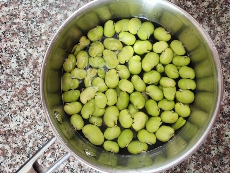 一位中年女性在明亮的现代厨房中操作不锈钢锅具,锅内正在煮沸的水中翻滚着绿色带壳鲜蚕豆,旁边放置玻璃碗盛有已剥壳的蚕豆,台面上有盐罐、橄榄油瓶和新鲜香草,体现健康烹饪氛围 家庭厨房中正在煮蚕豆的场景