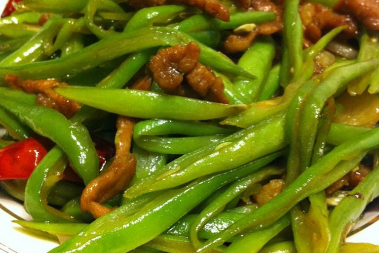 干煸长豆角成品特写：深绿色豆角段呈自然弯曲状，表面布满浅褐色虎皮褶皱，部分边缘微焦卷曲，盘底少油光亮，撒有少量干辣椒段与蒜末