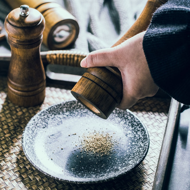 一位中年女性在明亮厨房中手持木质胡椒研磨器,正在将现磨黑胡椒撒入冒着热气的陶瓷炖盅内,汤面漂浮着鸡肉与枸杞,桌上有生姜片和砂锅,展现健康饮食的生活方式 家庭厨房中使用胡椒研磨器为炖汤添加黑胡椒的场景