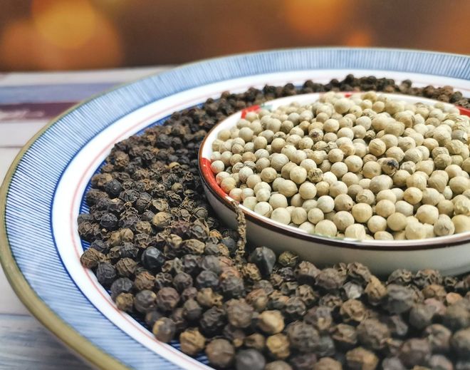 高清俯拍图:左侧为颗粒状黑胡椒,表面粗糙有褶皱,颜色深褐近黑;右侧为白胡椒,呈灰白色或浅米色,表面较光滑,形状规则圆形。背景为木质砧板,旁边配有研磨瓶和量勺,体现厨房使用场景 白胡椒与黑胡椒实物对比图