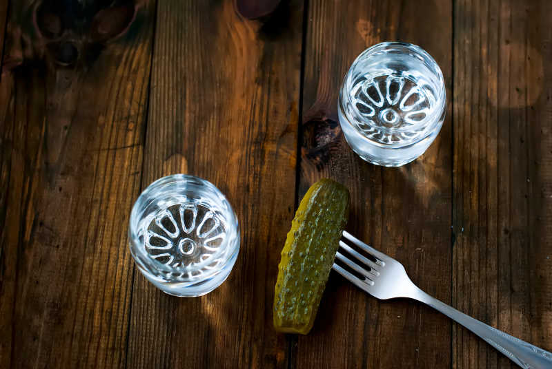 一张木质餐桌上摆放着一瓶透明伏特加酒、一块切开的黑麦面包、一碟切片的俄式小黄瓜,旁边还有酸奶油和新鲜莳萝点缀,整体呈现浓郁的东欧乡村风情 俄式小黄瓜搭配伏特加与黑麦面包的餐桌摆盘