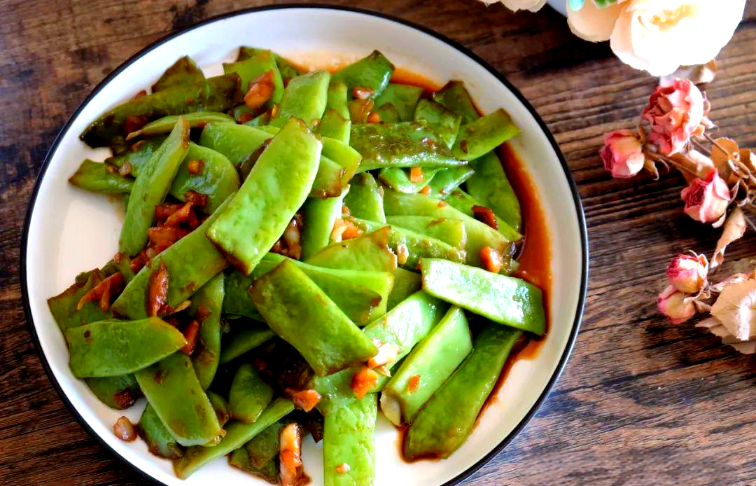 一盘刚出锅的清炒扁豆,色泽鲜绿油亮,配有少许蒜末和红色辣椒点缀,盛放在白色瓷盘中,背景为木质餐桌,整体风格清新自然 炒扁豆成品菜肴展示