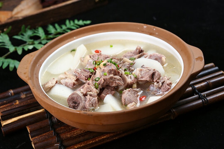 一个黑色砂锅正放在炉灶上慢炖,锅盖半开,可见内部翻滚的乳白色汤汁,羊肉块与白萝卜清晰可见,蒸汽袅袅升起,周围点缀着葱花和香菜 炖锅中正在慢炖的羊肉萝卜汤