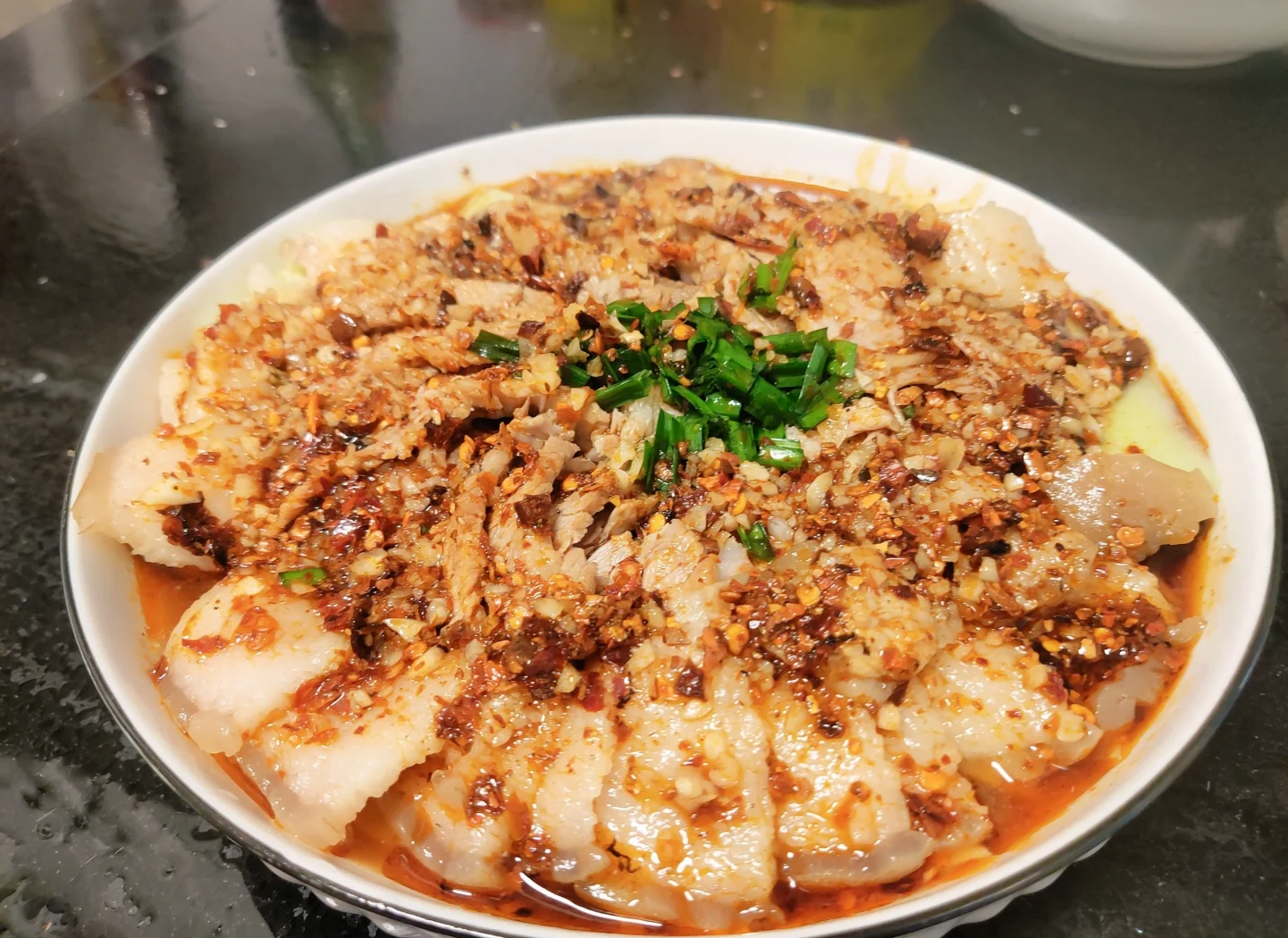 一盘红油亮泽的蒜泥白肉摆放在青瓷盘中，旁边配有蘸碟与筷子