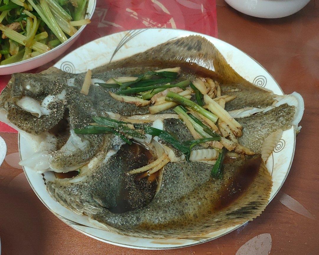 一位厨师正在厨房中将处理好的多宝鱼放入蒸锅，旁边摆放着姜片、葱段和酱油调料，准备进行清蒸烹饪