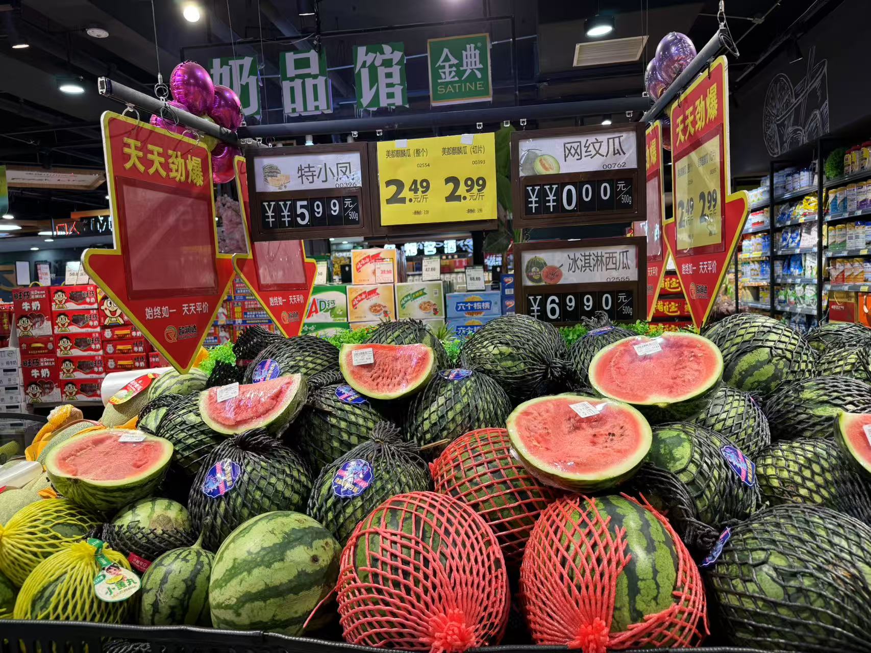 超市生鲜区的货架上整齐摆放着多个标有‘无籽西瓜’标签的圆形西瓜,表面光滑带有深绿条纹,部分已切开展示红色果肉,旁边有价格牌和促销标识,环境明亮整洁 超市货架上的无籽西瓜展示