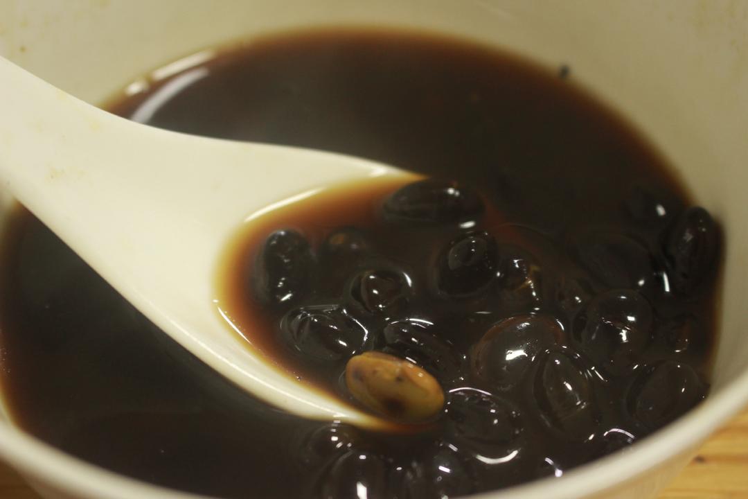 一碗刚煮好的黑豆黄豆水,液体呈淡褐色略带红晕,表面有轻微油膜,碗边配有滤网和勺子,背景为厨房灶台,锅中仍有余热蒸汽升腾 黑豆黄豆煮水成品图