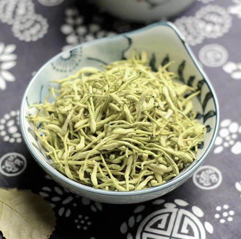 两组并列摆放的中药材:左侧为干燥的野菊花,花朵较小,呈深黄色,花瓣细长密集;右侧为干燥的金银花,花蕾呈棒状,初开者色黄白相间,未开放者表面覆有细柔毛。背景为木质茶盘,旁边配有玻璃量勺和透明玻璃杯,展示冲泡前状态 野菊花与金银花干品对比图