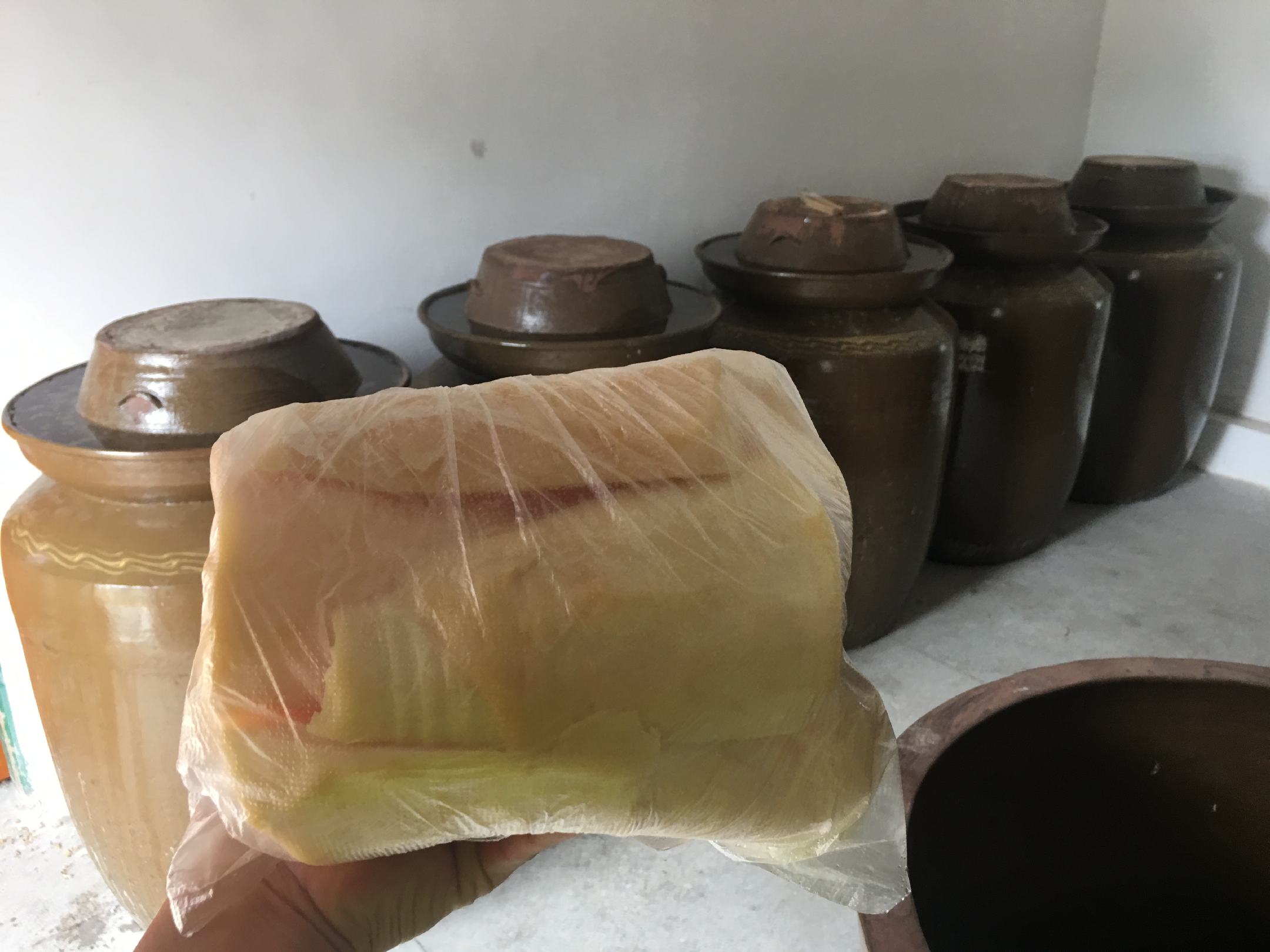 一个传统的陶制酸菜缸被放置在农家院角落,缸口覆盖着白色棉布,上面压着一块圆形青石,周围散落着几片掉落的白菜叶,背景可见木门与土墙 酸菜缸压石封存场景