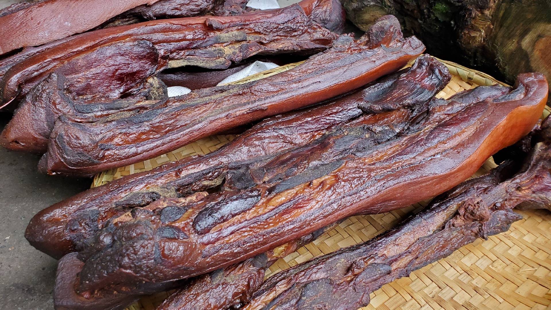 新鲜紫红色红菜薹整齐摆放在竹篮中,旁边放置一块切好的烟熏腊肉,背景为木质厨房台面,自然光线照射 红菜薹与腊肉原材料摆放