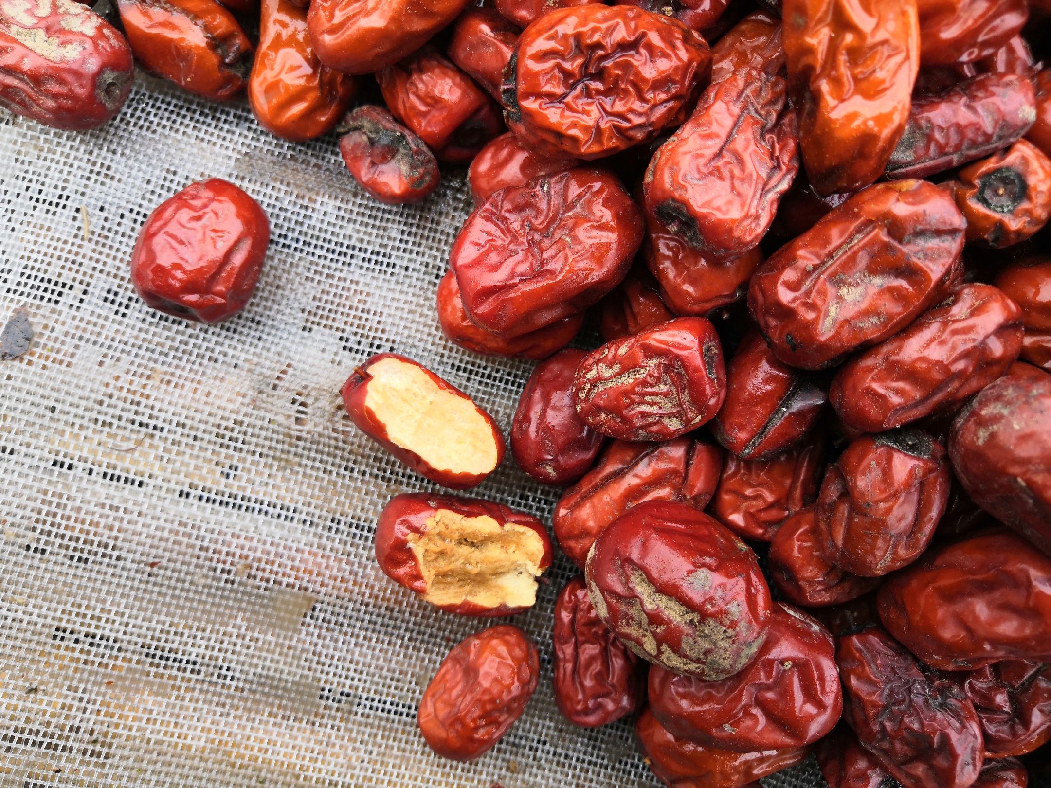 一颗饱满的金丝小枣,表面光滑呈深红色,光线照射下可见果肉透亮,部分果子已被掰开,露出内部晶莹的金黄色糖丝结构,背景为自然田园环境或晾晒场地 金丝小枣特写