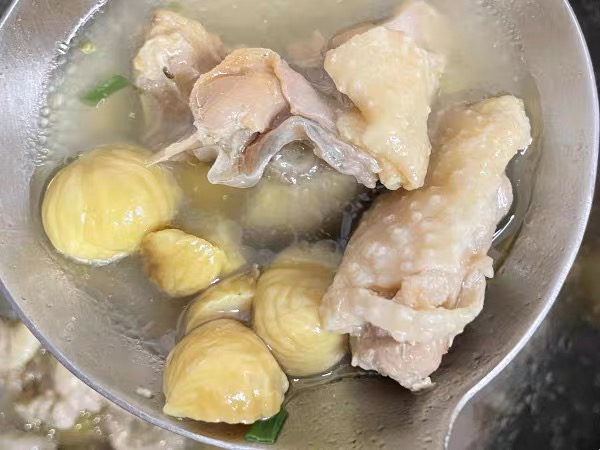 厨房台面上摆放一碗煮熟剥壳的金黄色板栗仁,旁边是一锅正在炖煮的鸡汤,汤中有整颗栗子和鸡块,配以胡萝卜片和香菜点缀,整体呈现温暖家常氛围 家庭烹饪板栗菜肴场景
