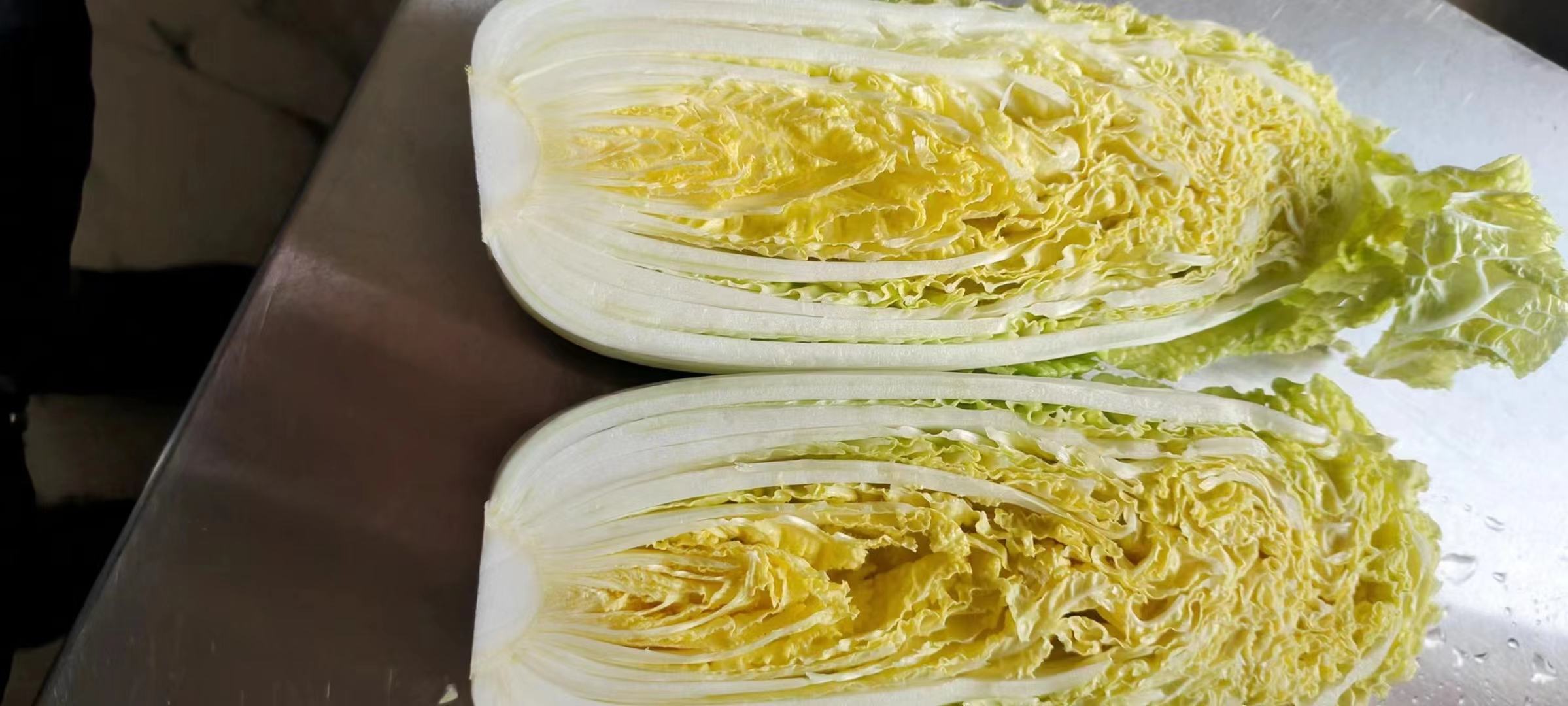 高清特写:娃娃菜采用纵向剖开+纤维走向轻推切法,切出厚度均一的1.5mm薄片,外叶完整保留用于装饰,刀具锋利无锯齿 娃娃菜双刀分切特写:砧板上整齐排列着纵向剖开的娃娃菜,外层嫩黄叶片完整剥离,菜心被切成均匀1.5mm厚的薄片,刀锋锐利反光,背景为木质纹理