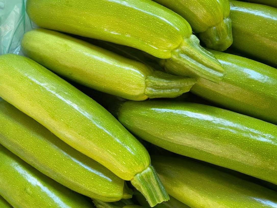 西葫芦与丝瓜超市货架并置实景图：左侧冷柜陈列带花西葫芦（浅绿短粗型，表面光亮，花鲜黄未萎）；右侧筐装丝瓜（墨绿细长型，表皮带浅棱与微绒，根部偶见残留雄花）