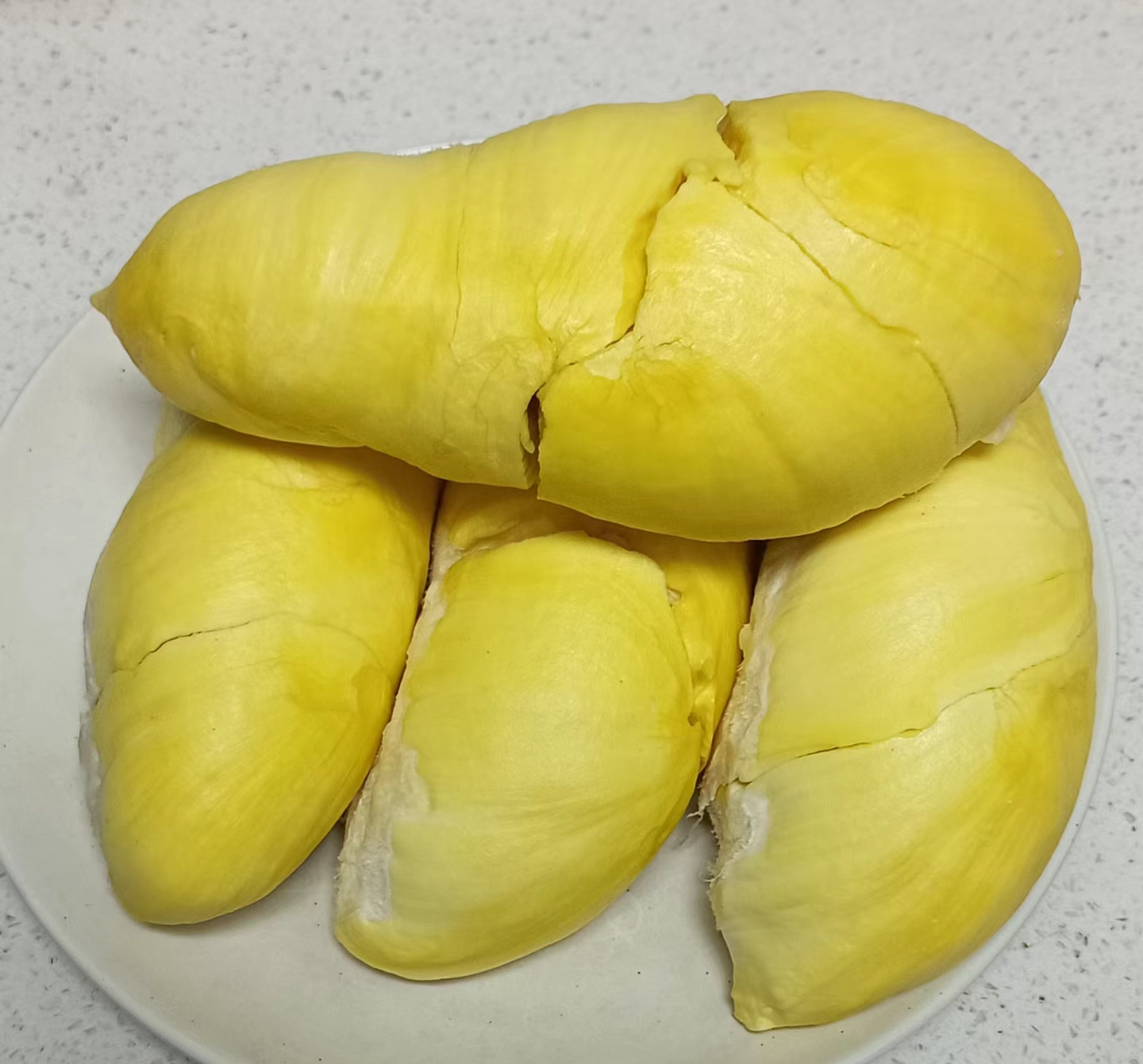 数块金枕头榴莲果肉整齐摆放在白色瓷盘中,果肉呈淡黄色,表面光滑有光泽,部分带有细微纤维纹理,盘边点缀一片新鲜柠檬叶,背景为模糊的室内用餐环境 金枕头榴莲果肉摆盘展示