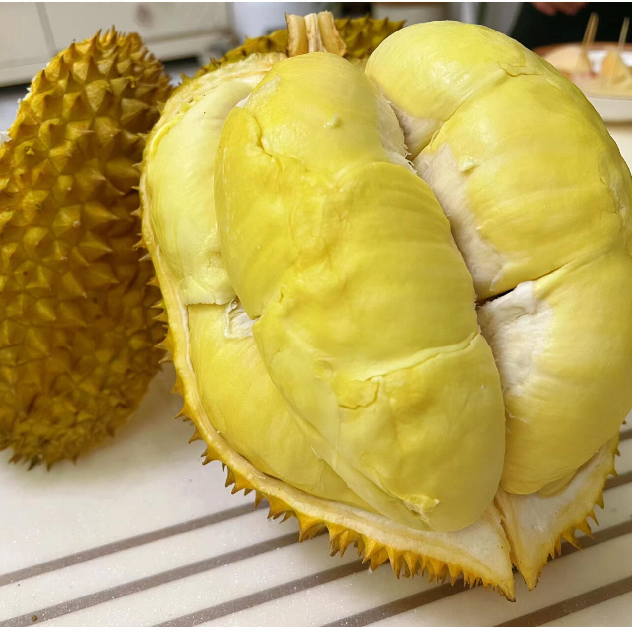 金枕头榴莲果肉特写拼盘,展示其厚实、完整、色泽均匀的果瓣,每块果肉清晰可见纤维结构,体现高品质新鲜度,适合用于美食宣传场景 多个切开的金枕头榴莲拼盘摆放在白色瓷盘中,果肉洁白如玉,排列整齐,背景为浅色厨房台面,营造美食氛围