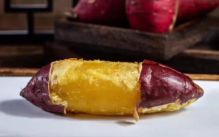 一根刚蒸好的红心红薯被横向切开,露出金红色的内瓤,热气袅袅上升,切面呈现出细腻的纤维结构,旁边摆放着白色瓷盘和银色小勺 蒸熟的红心红薯切开画面